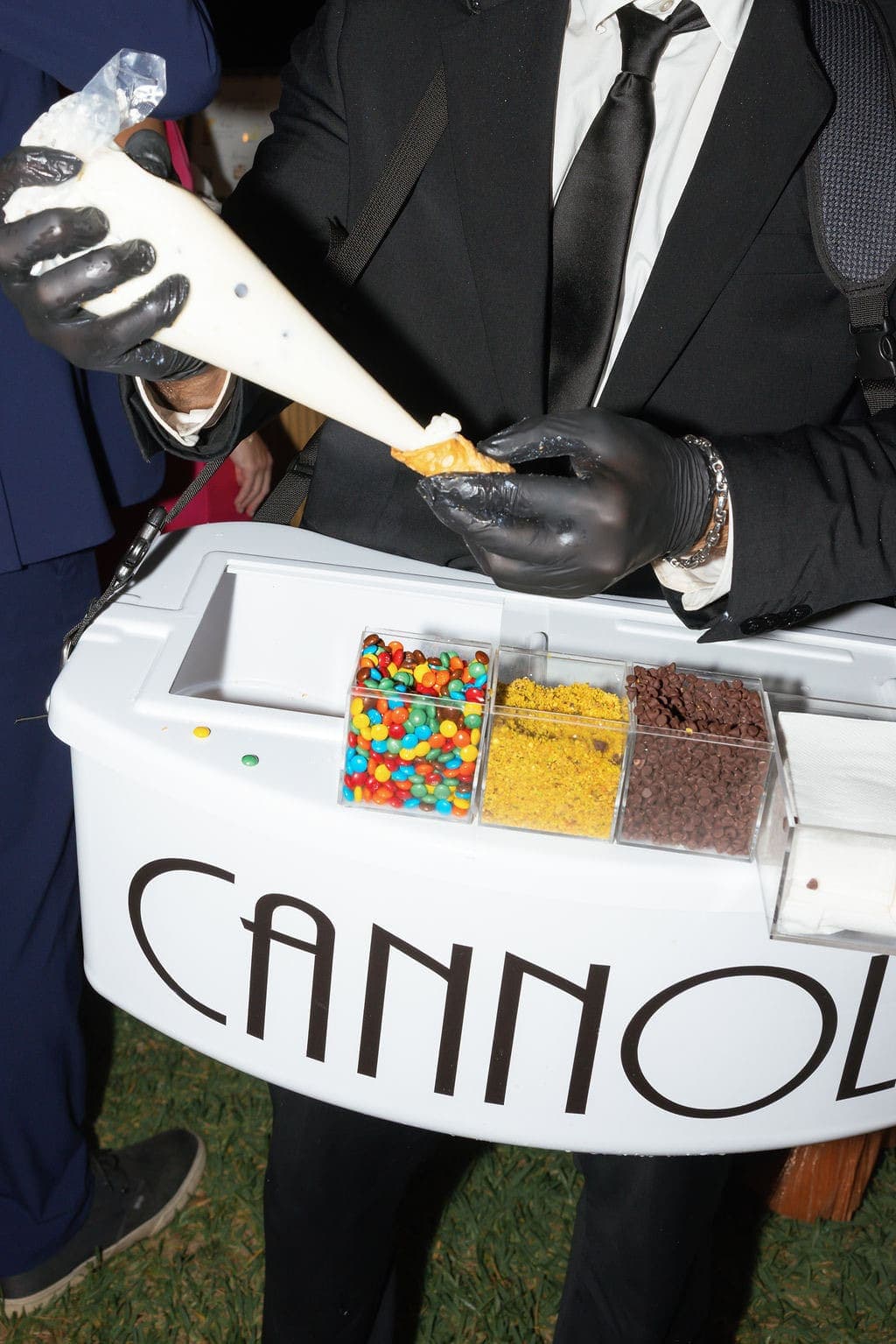 Tuxedoed cannoli server piping filling into a shell at an evening event