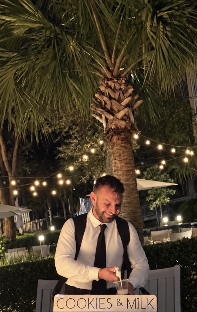 Bottoms Up Luxury attendant serving cookies and milk beneath string lights and palms at an evening event
